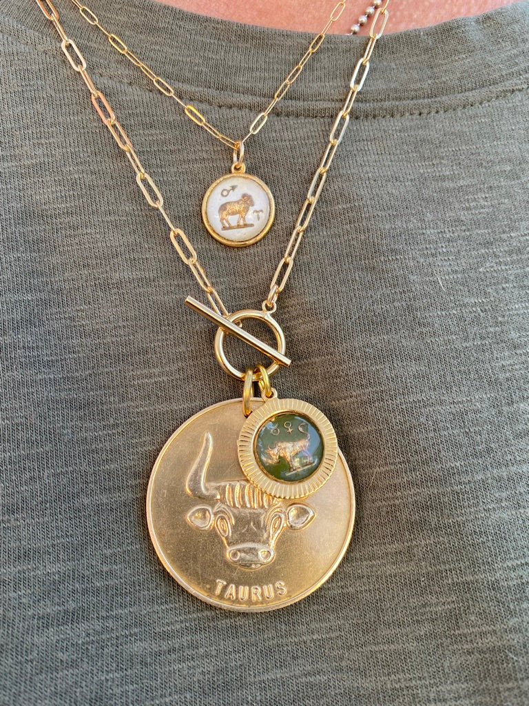Layered gold necklace look featuring a large Taurus zodiac coin pendant with bull design and a smaller round zodiac charm on gold paperclip chains with toggle clasp worn on a model in a gray top