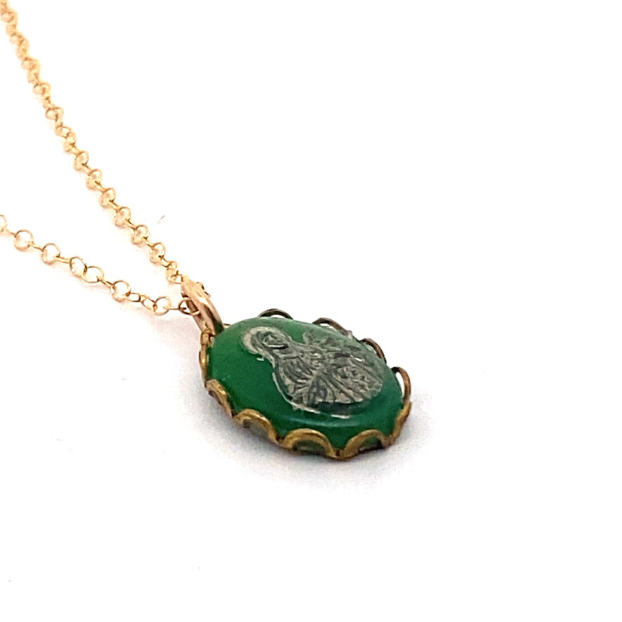 Side angle close-up of a vintage emerald green Virgin Mary cameo pendant with scalloped gold edge on a gold chain against a white background