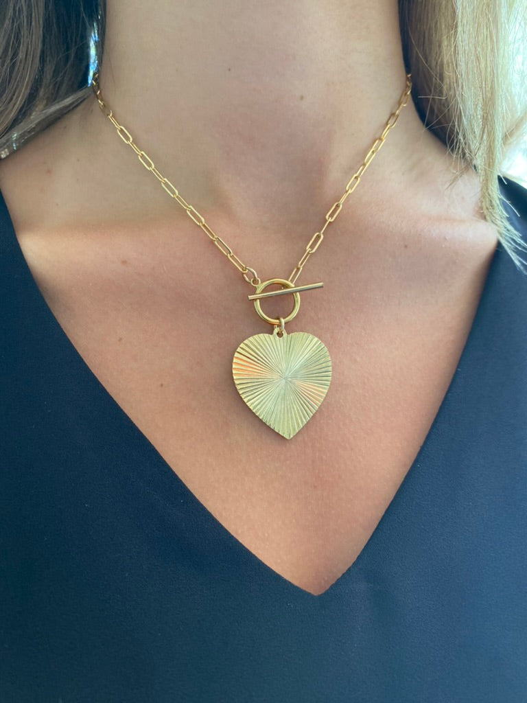 Gold ribbed heart-shaped pendant necklace with toggle clasp closure on a gold paperclip chain worn on a model in a dark top