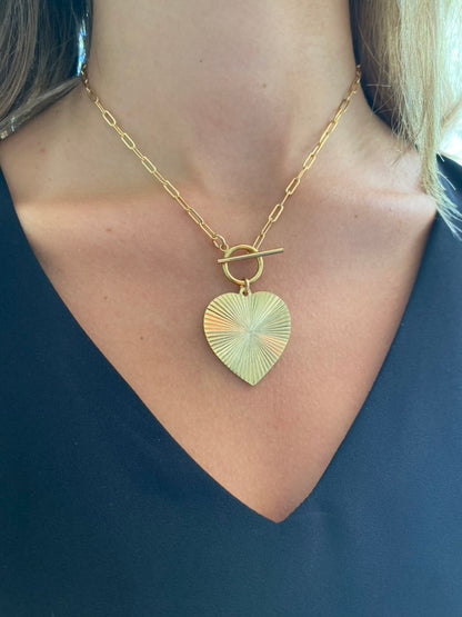 Gold ribbed heart-shaped pendant necklace with toggle clasp closure on a gold paperclip chain worn on a model in a dark top