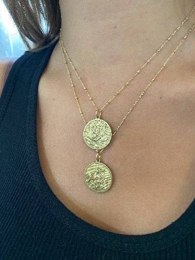 Two gold embossed birth month flower coin pendant necklaces layered on a model wearing a dark top lifestyle photo