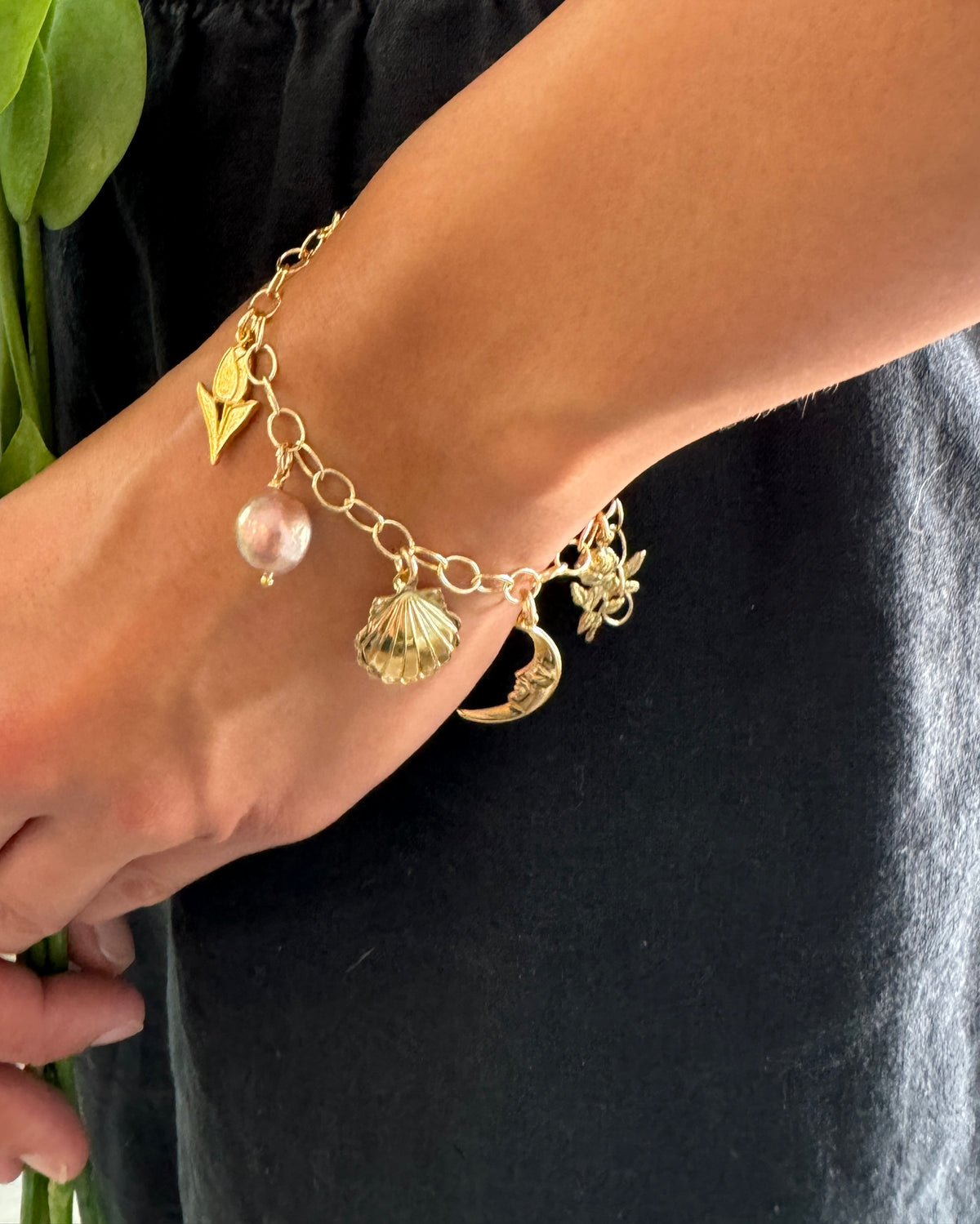 Gold charm bracelet with round link chain worn on a model featuring pearl, seashell, crescent moon, rose, cherub angel, and arrow charms against a dark background