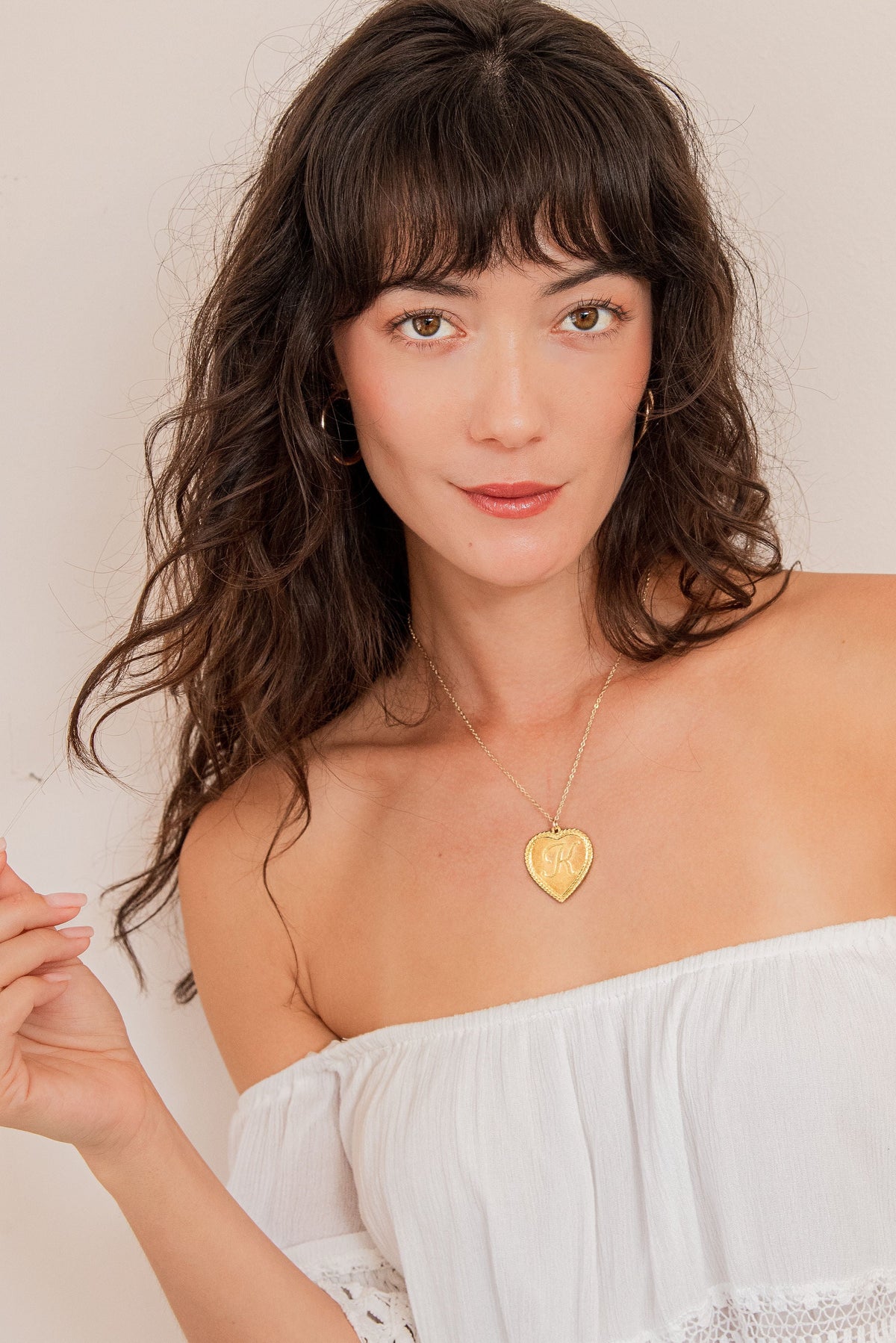 Woman in a white strapless top with a gold necklace with a heart pendant with the letter "K" in the middle 