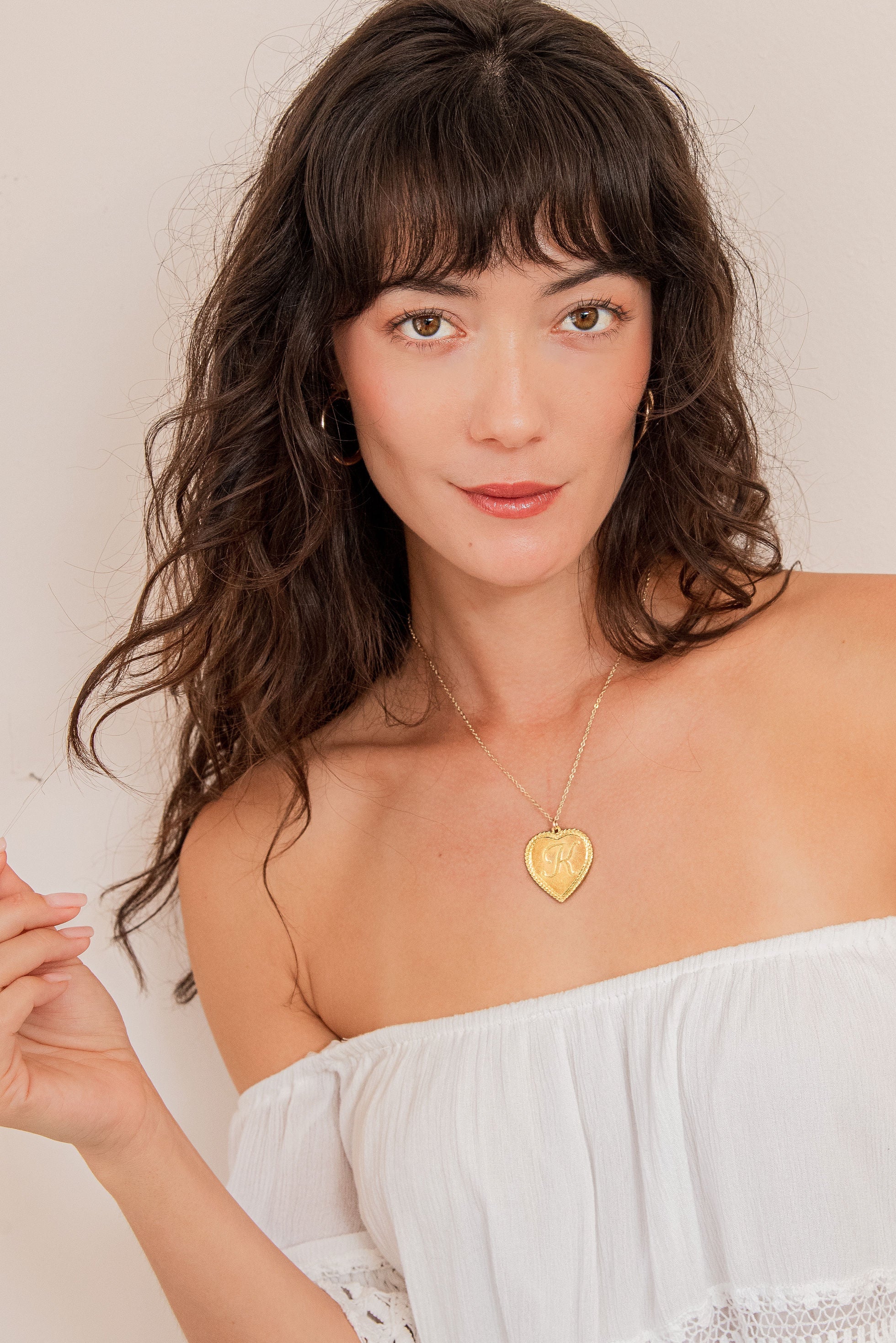 Woman in a white strapless top with a gold necklace with a heart pendant with the letter "K" in the middle 