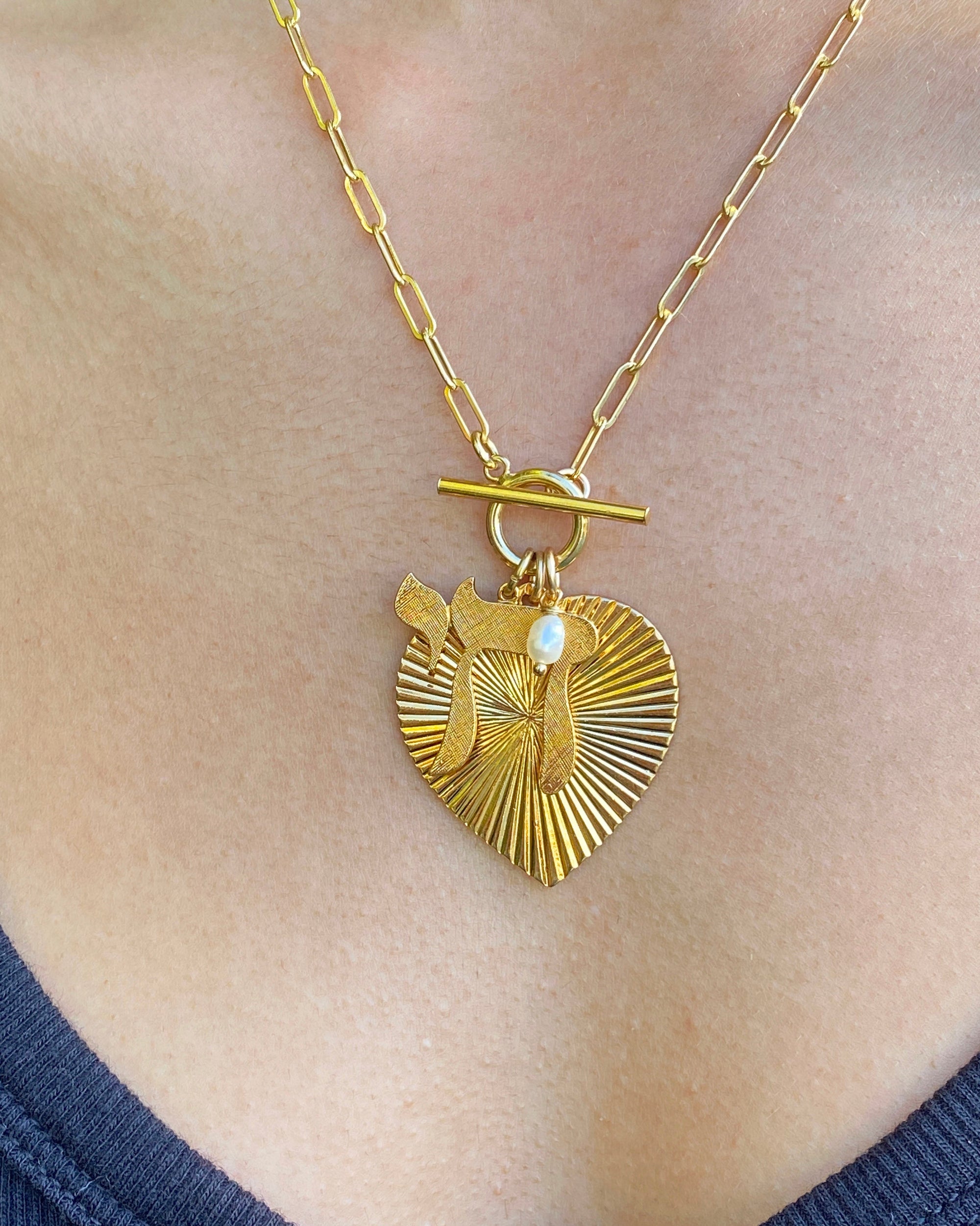 A gold ribbed heart pendant necklace with a Chai symbol and pearl accent on a toggle clasp paperclip chain, worn on a person's chest