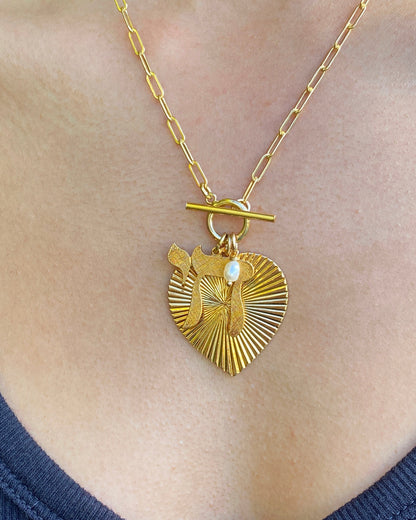 A gold ribbed heart pendant necklace with a Chai symbol and pearl accent on a toggle clasp paperclip chain, worn on a person's chest