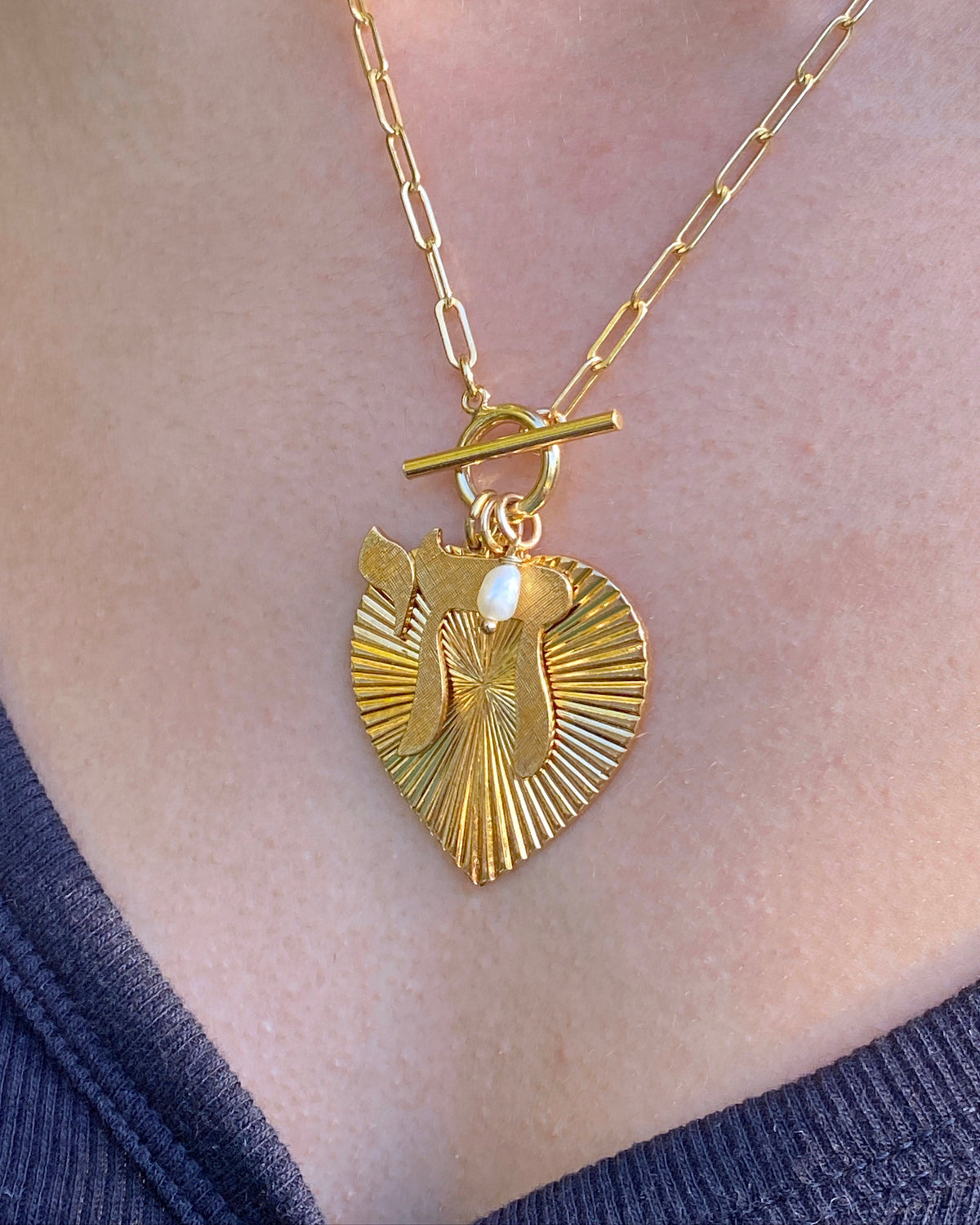 A gold-plated ribbed heart-shaped necklace with a textured chai charm and a fresh water pearl on a chain, displayed on a person's neck.