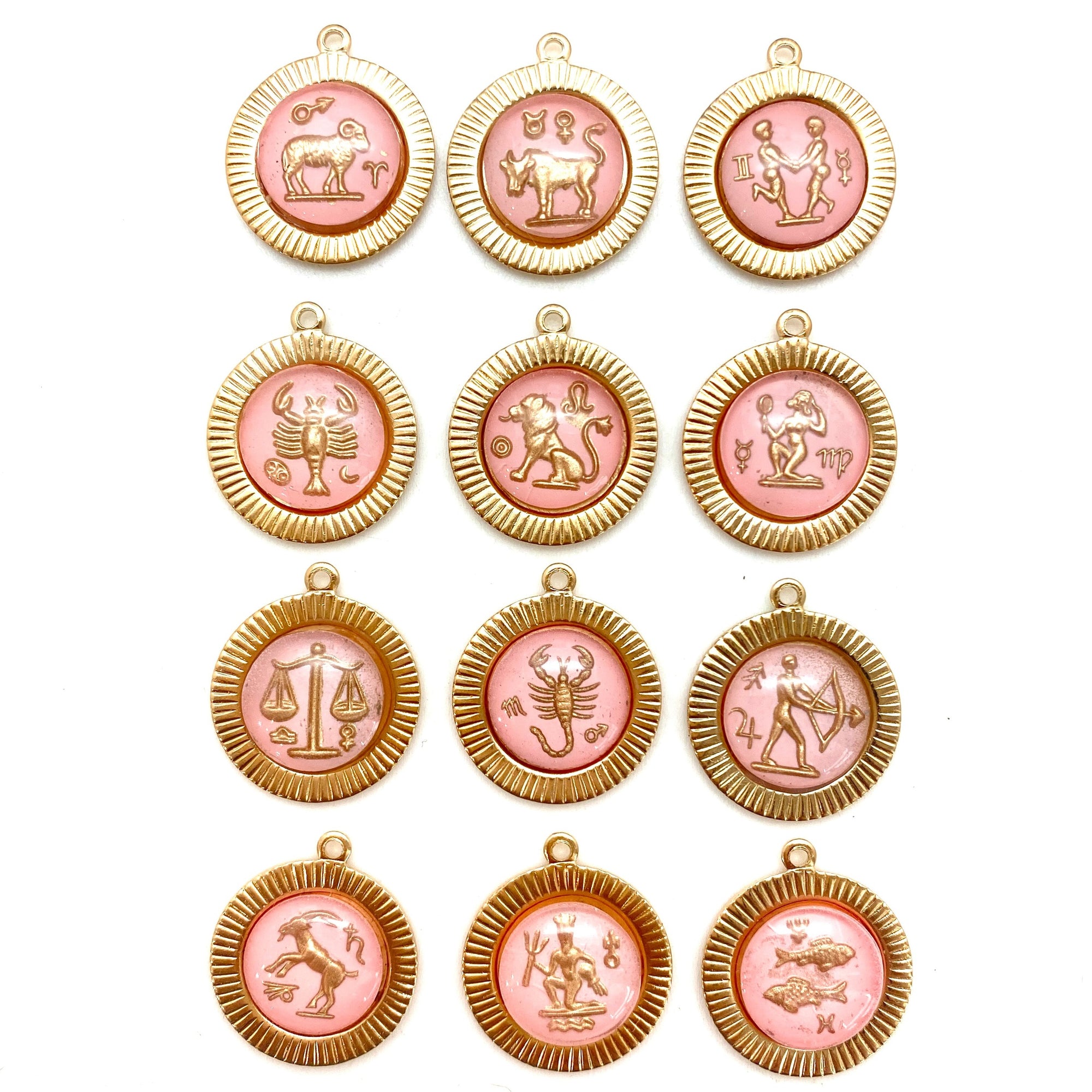 Collection of 12 pink enamel zodiac sign round pendants with gold sunburst ribbed frames arranged in a grid on a white background featuring Aries, Taurus, Gemini, Cancer, Leo, Virgo, Libra, Scorpio, Sagittarius, Capricorn, Aquarius, and Pisces with gold zodiac figure intaglio designs