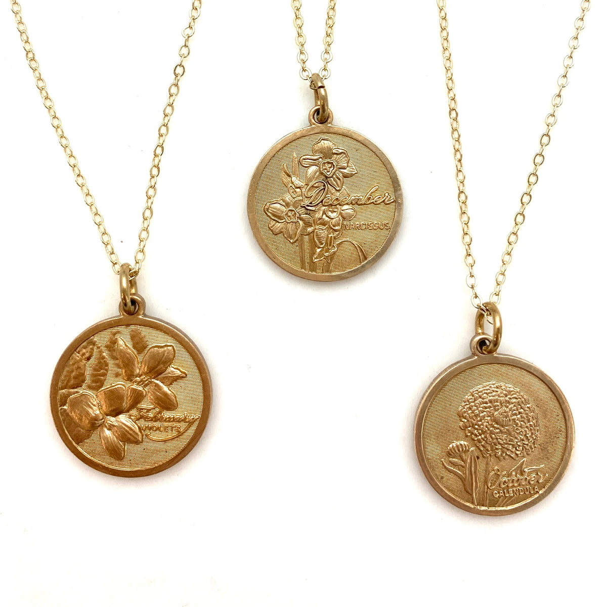 Three gold embossed birth month flower coin pendant necklaces on delicate gold chains featuring February violet December narcissus and October calendula on a white background