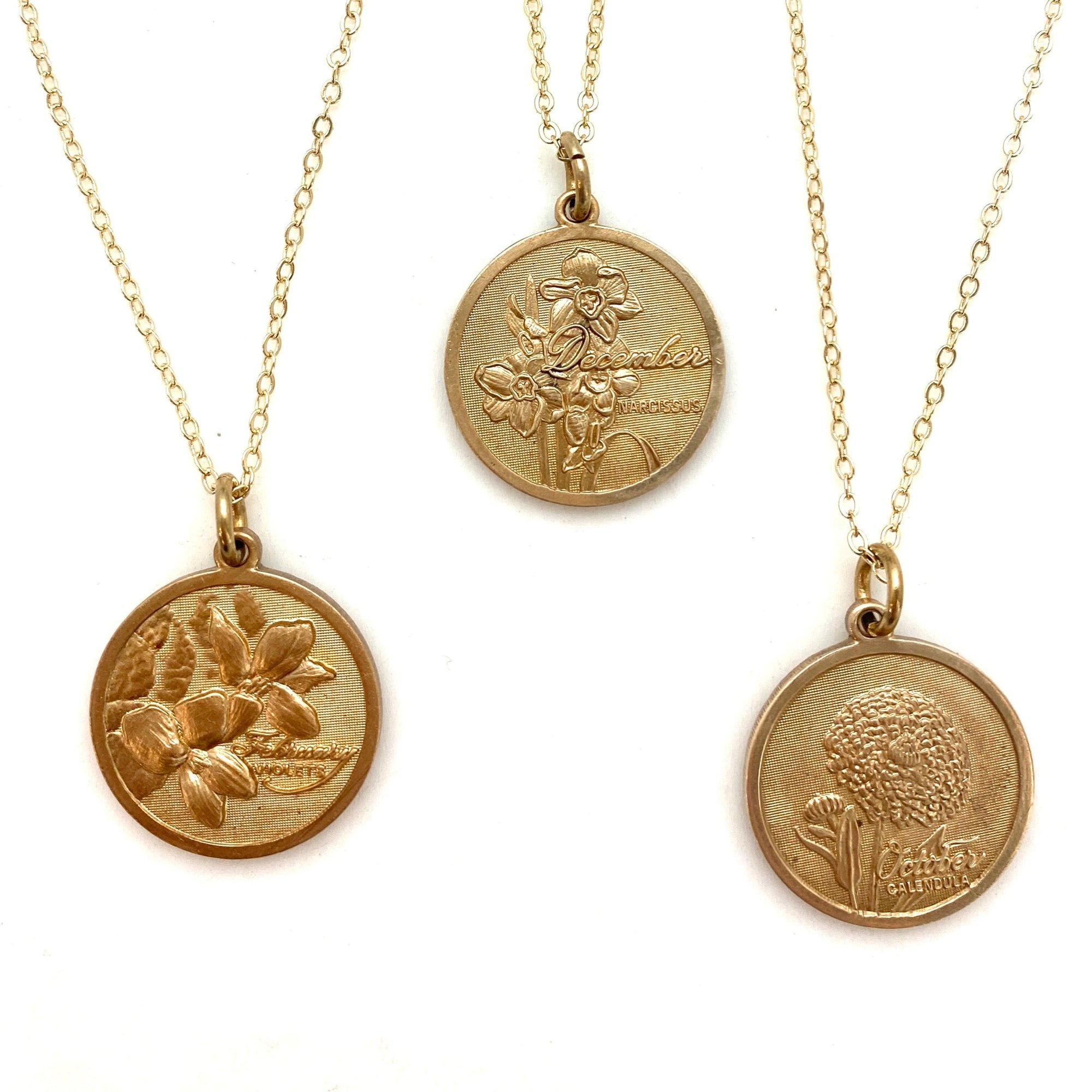 Three gold embossed birth month flower coin pendant necklaces on delicate gold chains featuring February violet December narcissus and October calendula on a white background