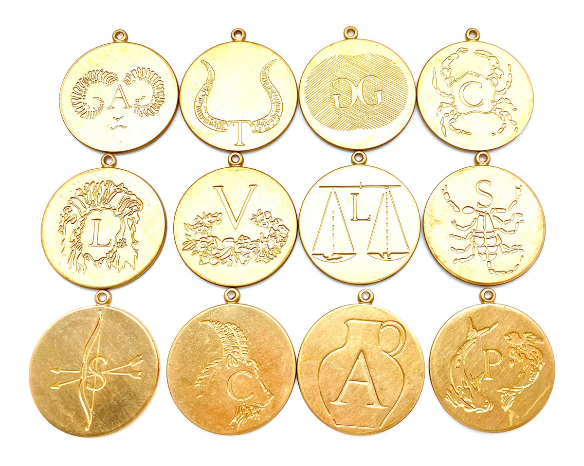 Collection of 12 gold engraved zodiac coin pendants arranged in a grid on a white background featuring Leo lion, Taurus bull, Gemini twins, Cancer crab, Leo, Virgo maiden, Libra scales, Scorpio scorpion, Sagittarius archer arrows, Capricorn sea goat, Aries ram, and Pisces fish with initial letter engravings