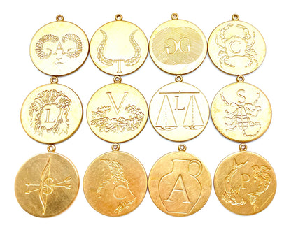 Collection of 12 gold engraved zodiac coin pendants arranged in a grid on a white background featuring Leo lion, Taurus bull, Gemini twins, Cancer crab, Leo, Virgo maiden, Libra scales, Scorpio scorpion, Sagittarius archer arrows, Capricorn sea goat, Aries ram, and Pisces fish with initial letter engravings