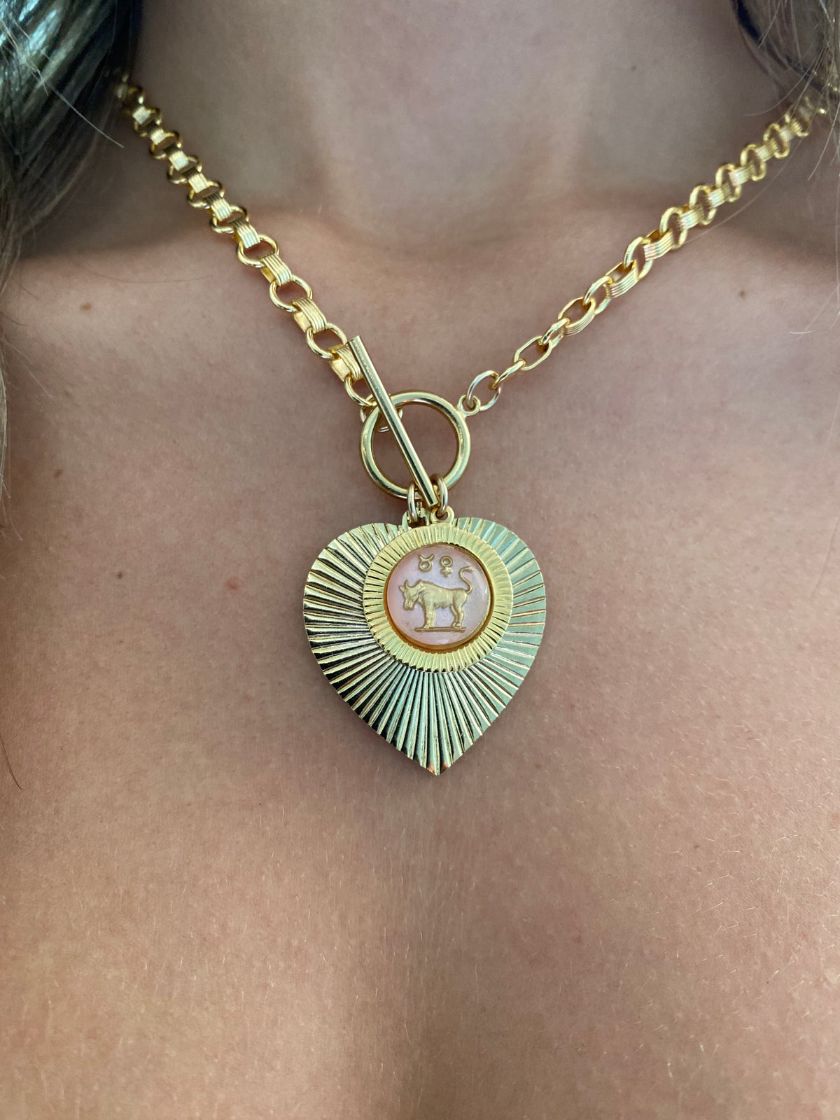 Gold and green patina heart-shaped sunburst pendant necklace with a pink vintage glass Taurus bull intaglio cameo on a gold toggle chain worn on a model close-up