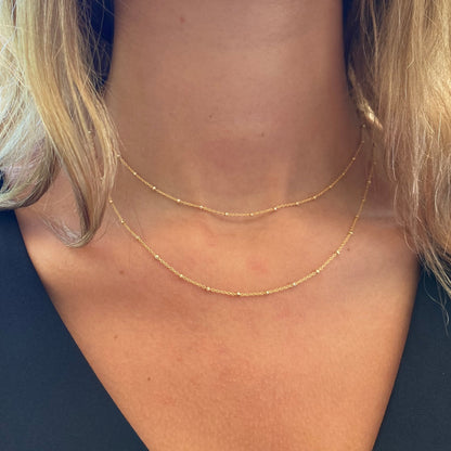 Layered gold satellite beaded chain necklaces worn on a model showing two delicate chains at different lengths against the neckline