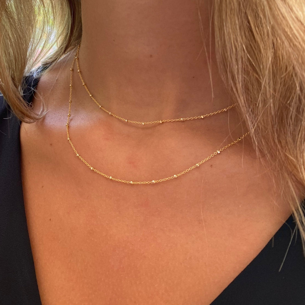 Layered gold satellite beaded chain necklaces worn on a model showing two delicate chains at different lengths in a close-up angled view