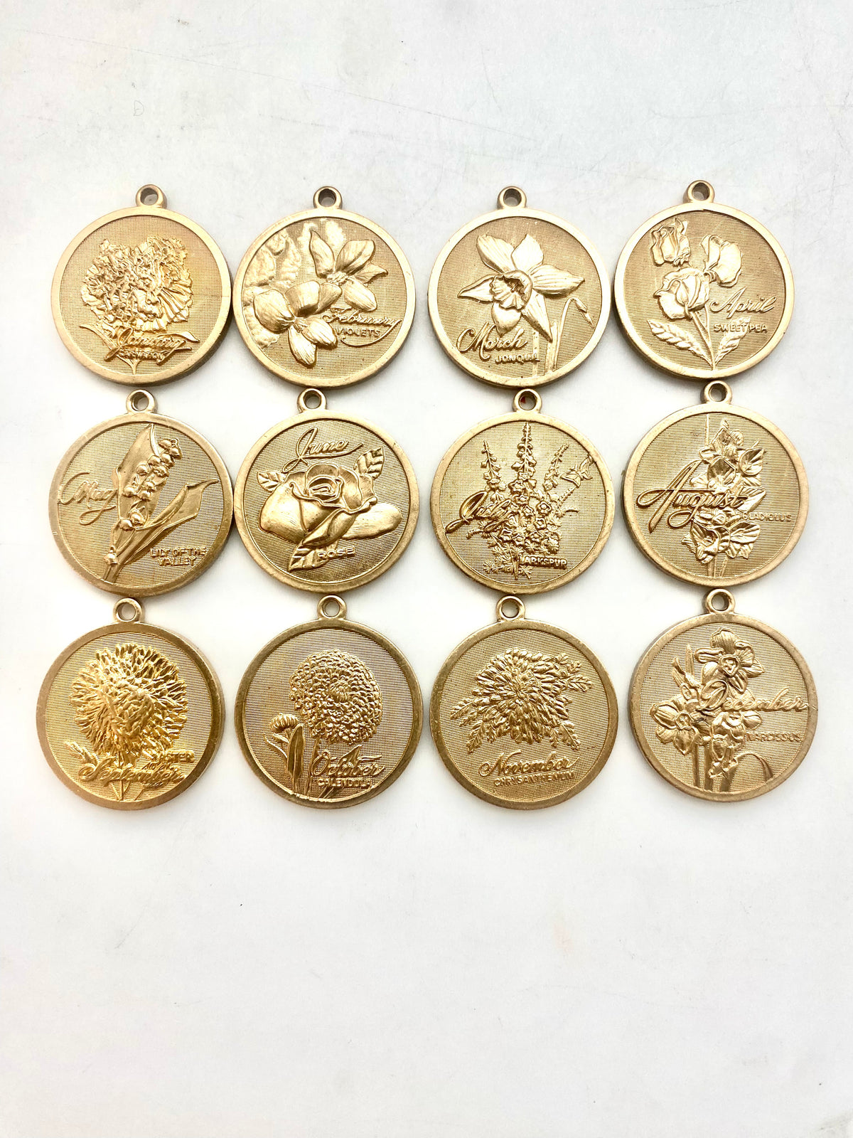 Collection of twelve gold embossed birth month flower coin pendant charms featuring all twelve months with floral designs arranged in a grid on a white background