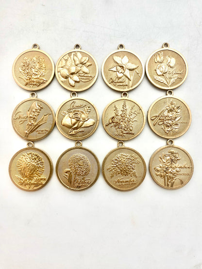 Collection of twelve gold embossed birth month flower coin pendant charms featuring all twelve months with floral designs arranged in a grid on a white background
