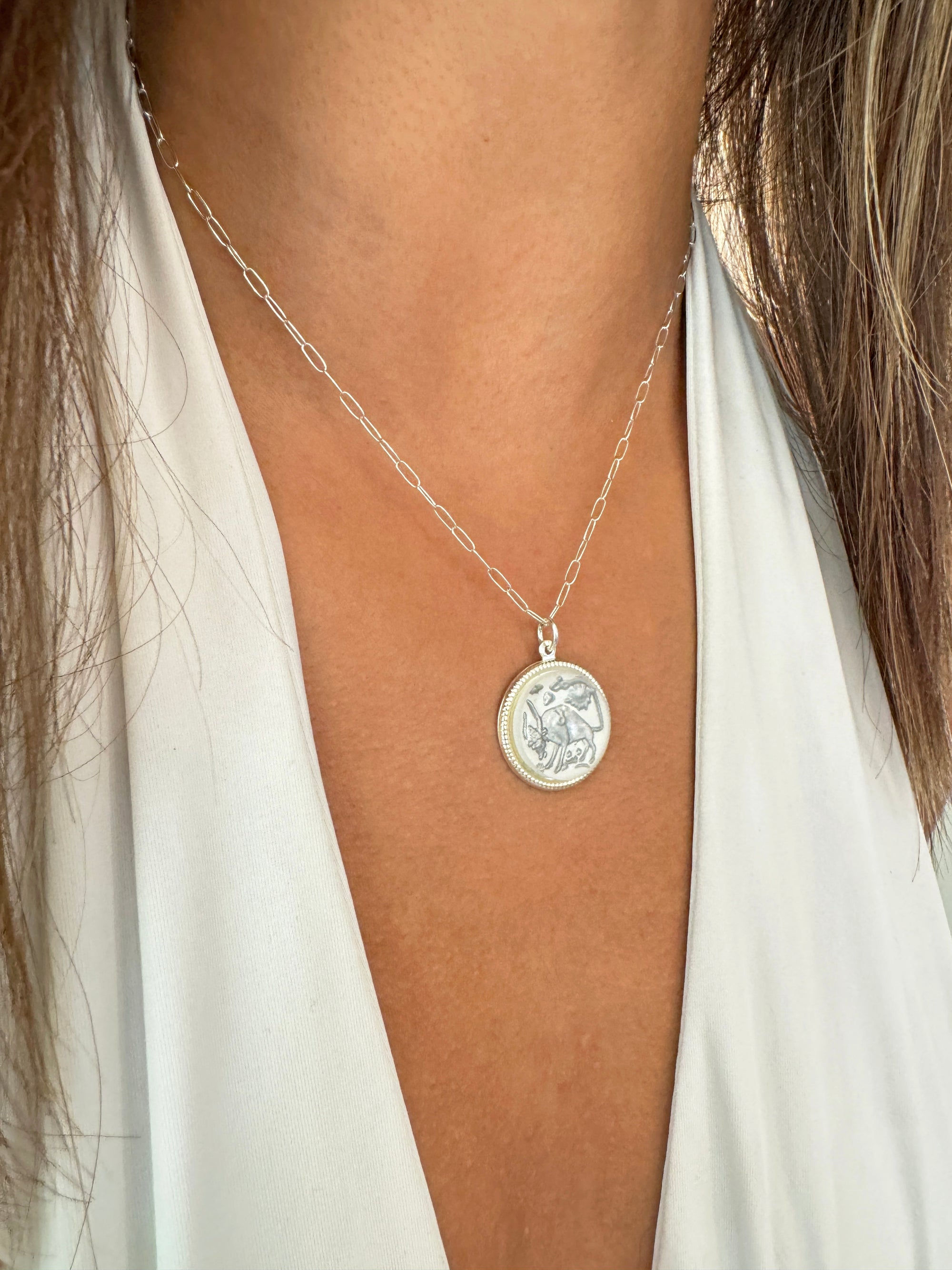 Close-up of a silver round Taurus zodiac coin pendant with bull design on a silver paperclip chain worn on a model in a white top