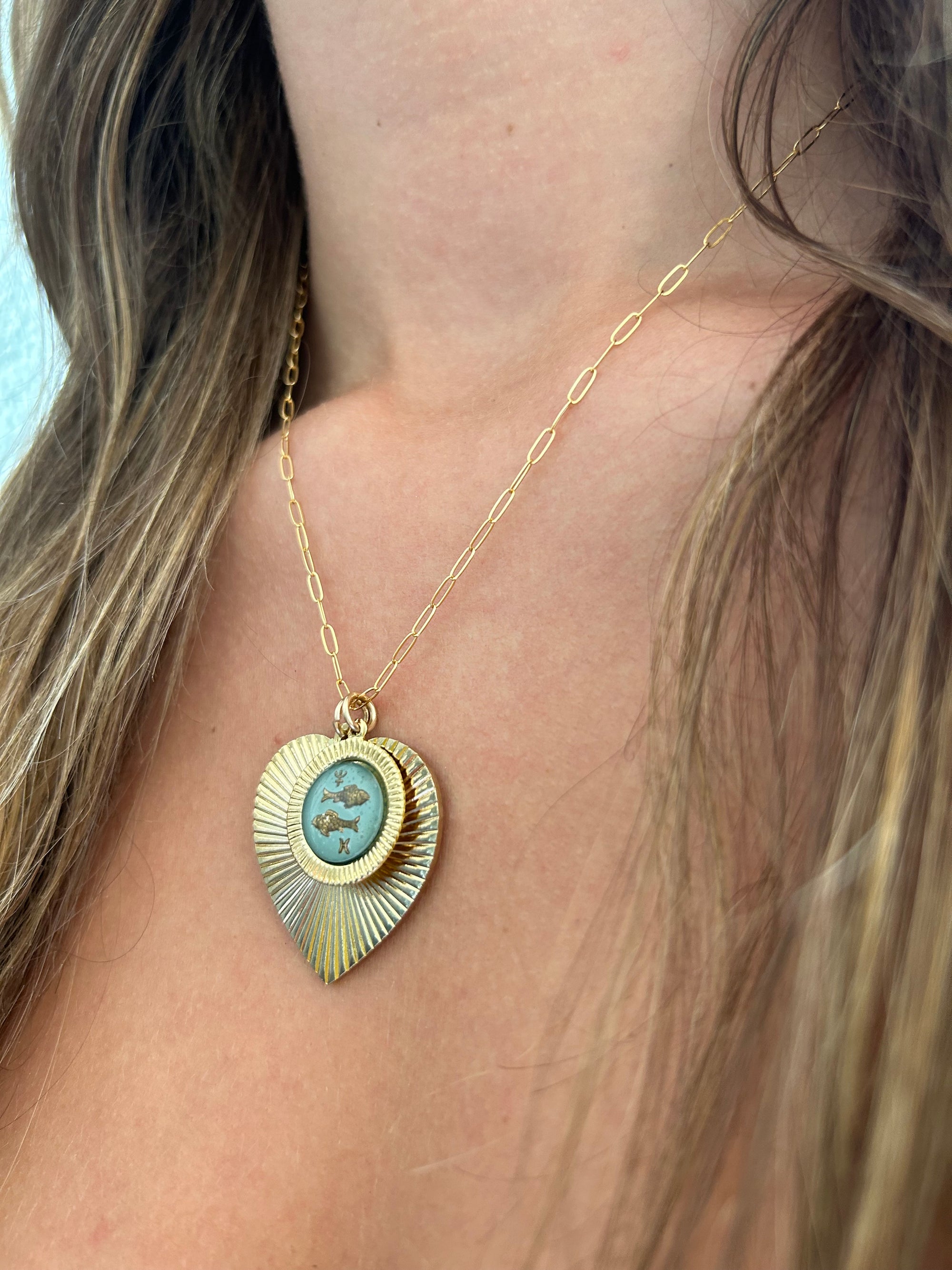 Gold heart-shaped sunburst pendant necklace with a light blue vintage glass Pisces fish intaglio cameo on a gold paperclip chain worn on a model close-up lifestyle photo