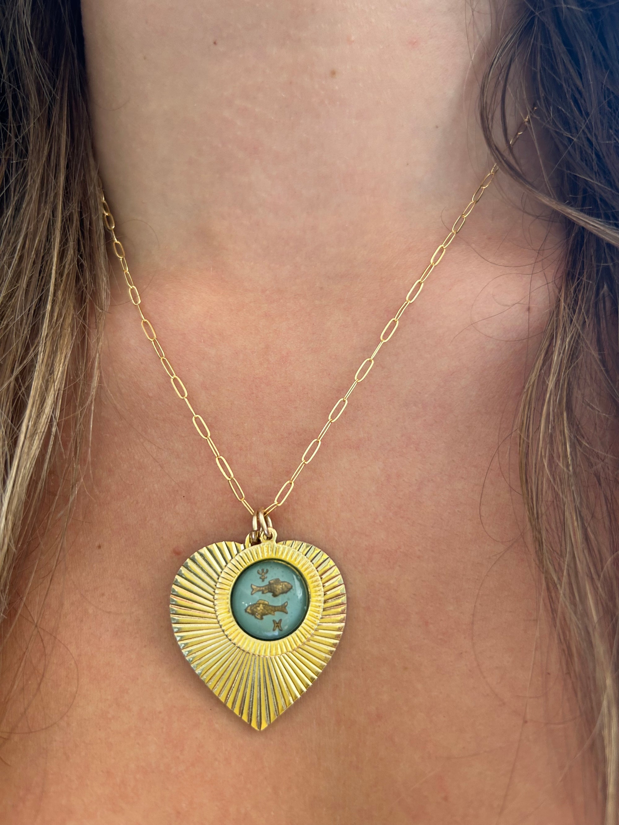 Gold heart-shaped sunburst pendant necklace with a light blue vintage glass Pisces fish intaglio cameo on a gold paperclip chain worn on a model close-up