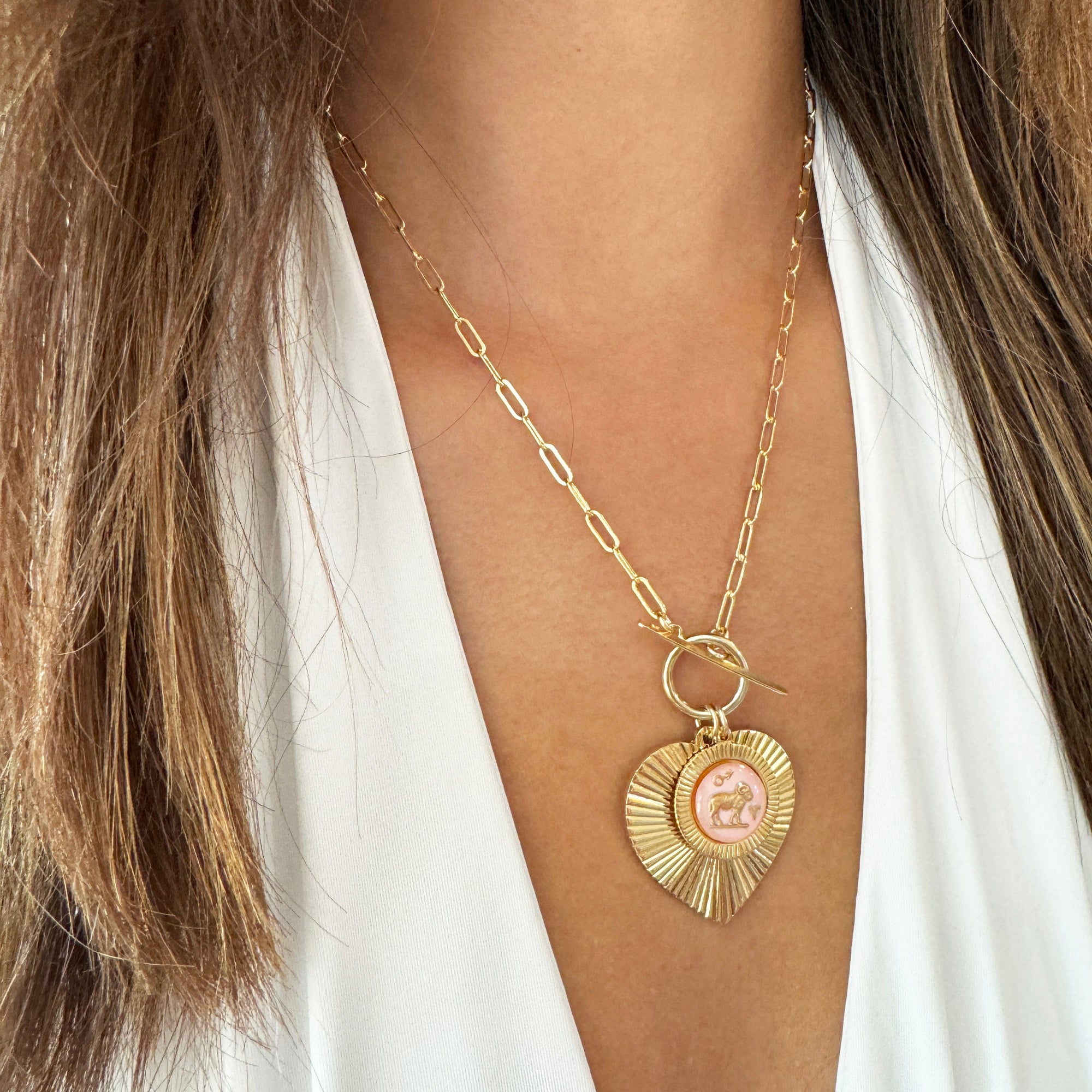 Gold ribbed heart-shaped pendant necklace with an Aries zodiac ram design in pink enamel center with toggle clasp on a gold paperclip chain worn on a model in a white top showing full necklace drape