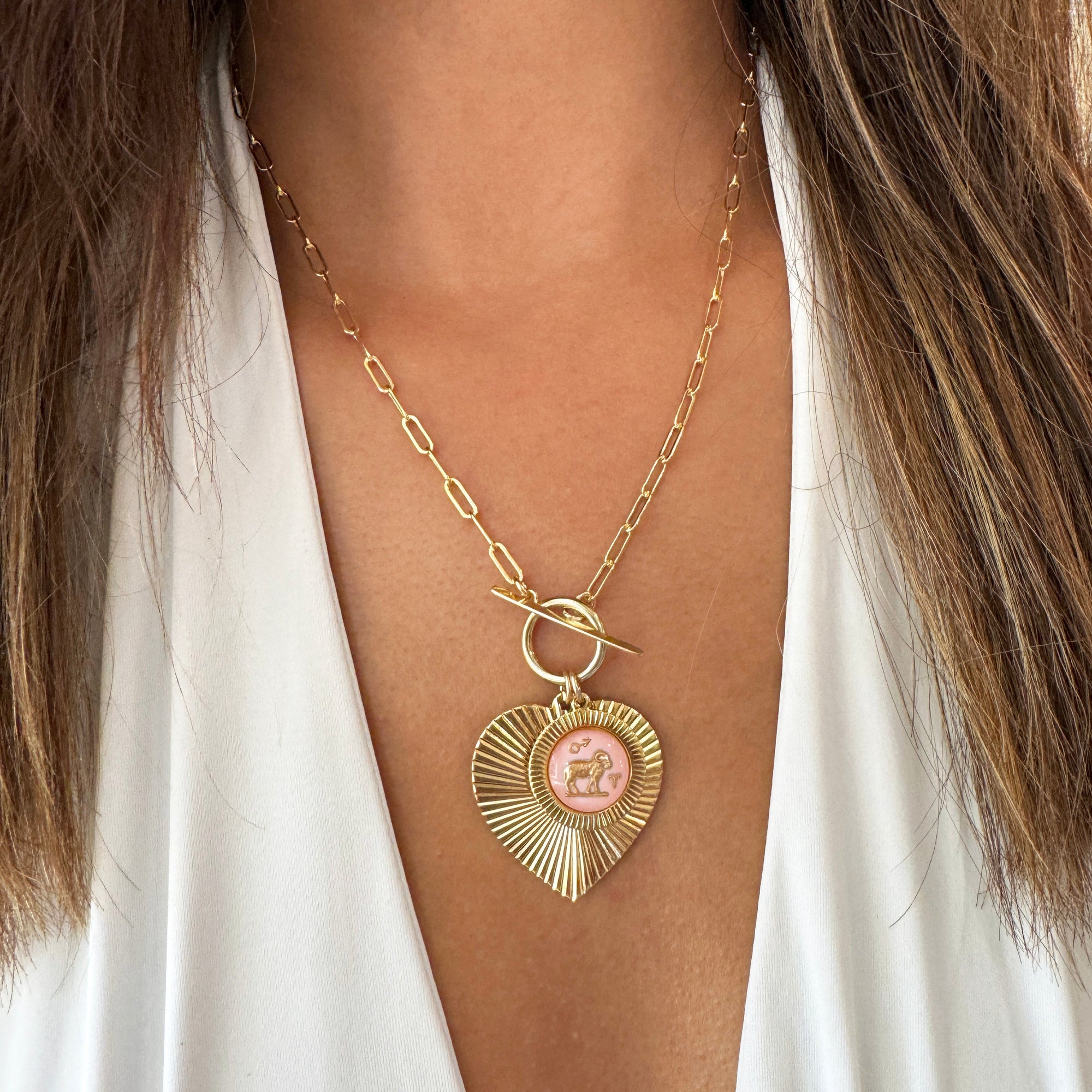 Gold ribbed heart-shaped pendant necklace with an Aries zodiac ram design in pink enamel center with toggle clasp on a gold paperclip chain worn on a model in a white top