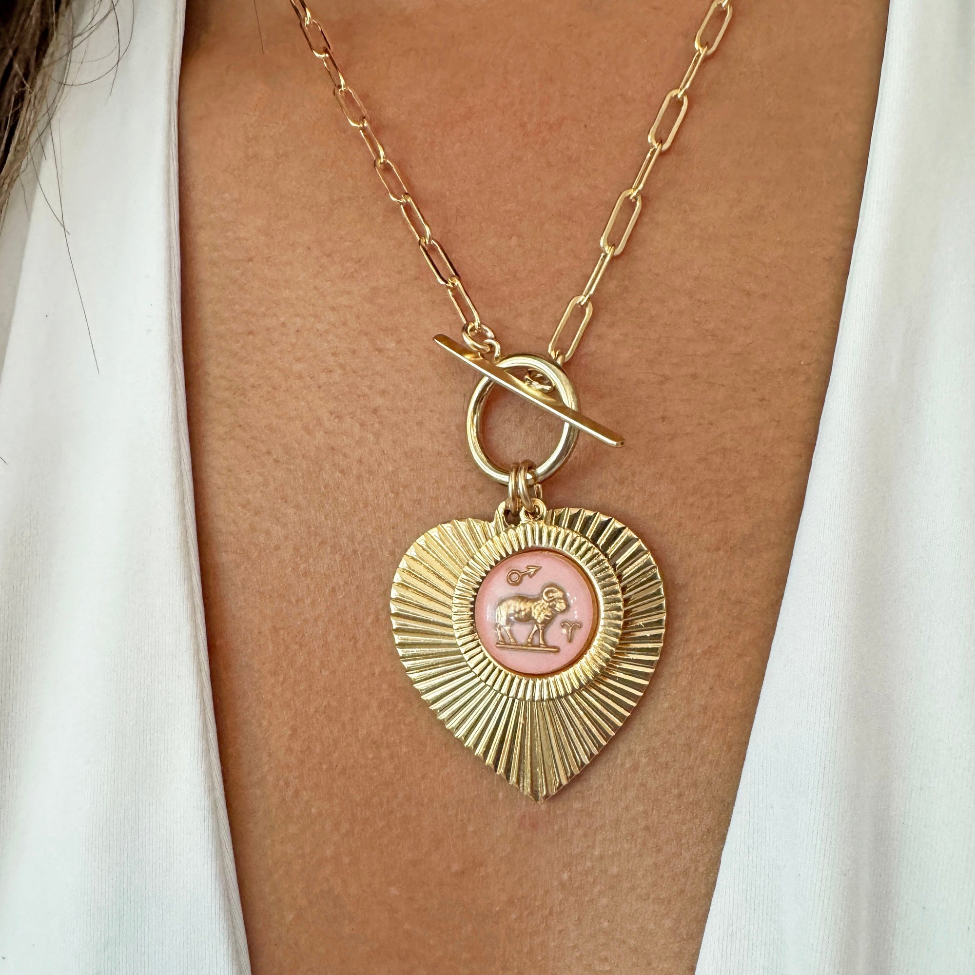 Gold ribbed heart-shaped pendant necklace with an Aries zodiac ram design in pink enamel center with toggle clasp on a gold paperclip chain worn on a model in a white top
