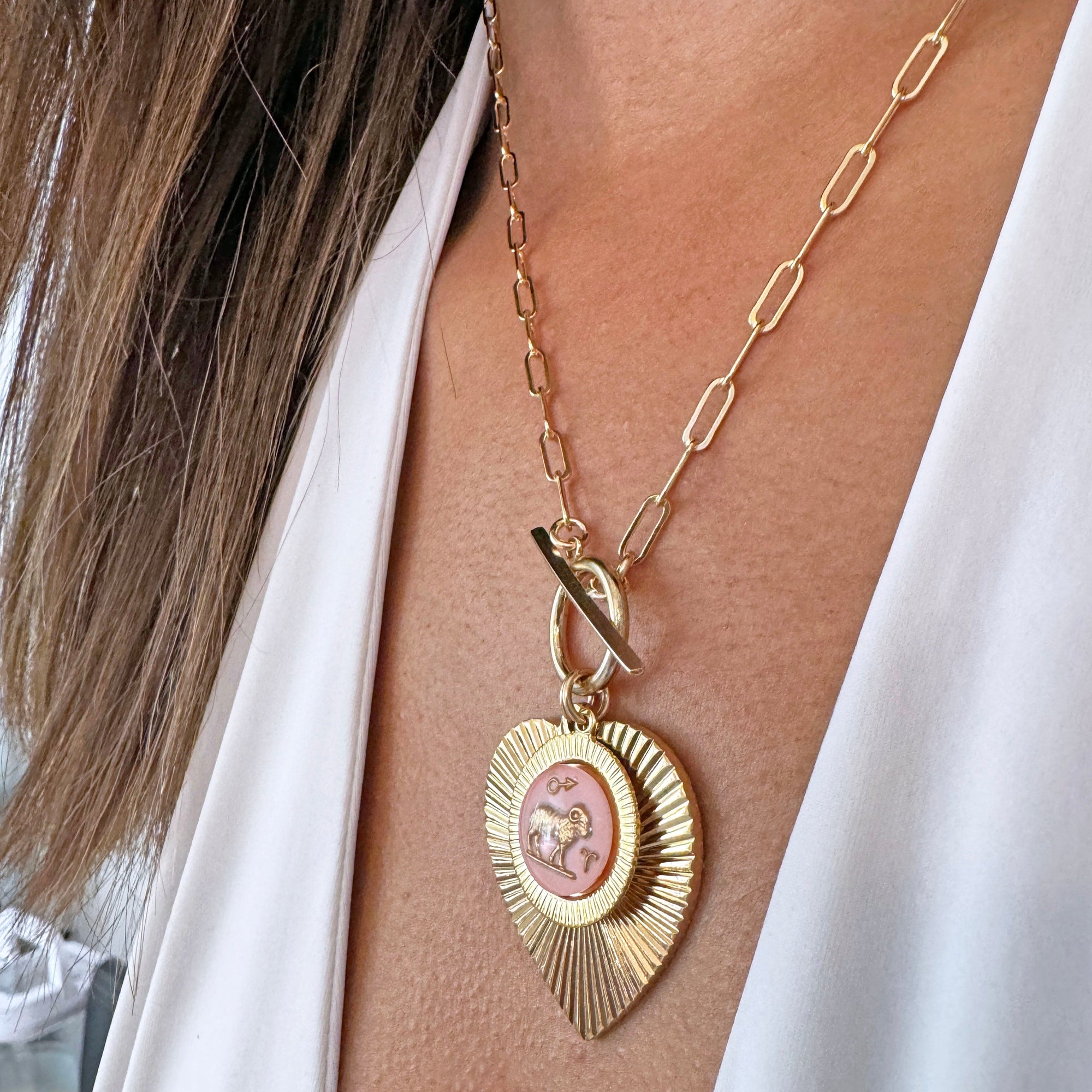 Gold ribbed heart-shaped pendant necklace with an Aries zodiac ram design in pink enamel center with toggle clasp on a gold paperclip chain worn on a model in a white top