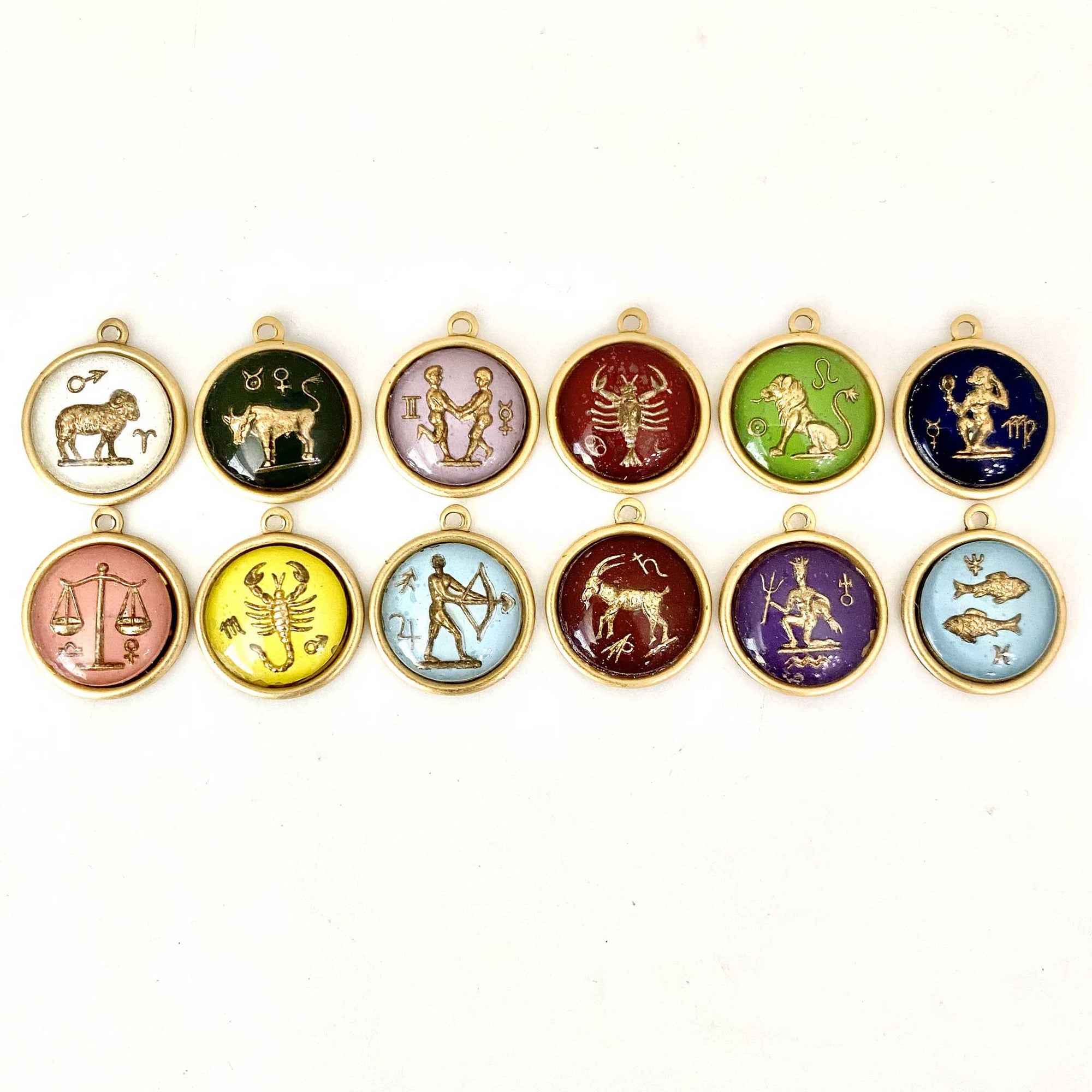 Collection of 12 small vintage German glass zodiac sign round pendants in assorted colors arranged in two rows on a white background featuring all zodiac signs including Aries, Taurus, Gemini, Cancer, Leo, Virgo, Libra, Scorpio, Sagittarius, Capricorn, Aquarius, and Pisces intaglio cameos in gold-framed settings