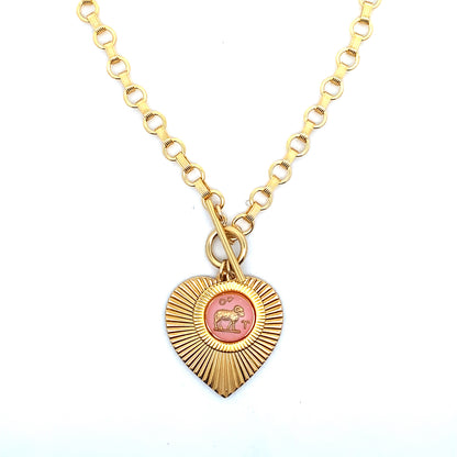 A gold ribbed heart-shaped pendant necklace with an Aries zodiac ram design in pink enamel center with toggle clasp closure on a gold round link chain against a white background