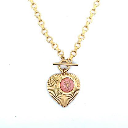 A gold ribbed heart-shaped pendant necklace with a Scorpio zodiac scorpion design in pink enamel center with toggle clasp closure on a gold round link chain against a white background