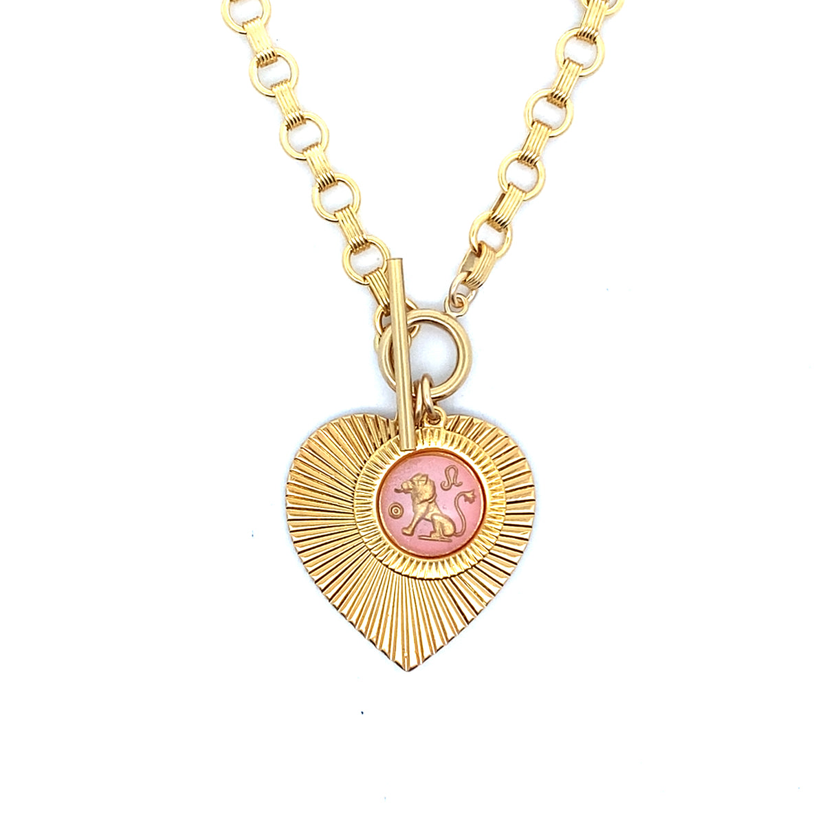 A gold ribbed heart-shaped pendant necklace with a Leo zodiac lion design in pink enamel center with toggle clasp closure on a gold round link chain against a white background