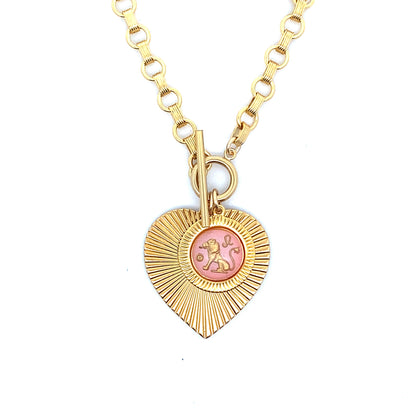 A gold ribbed heart-shaped pendant necklace with a Leo zodiac lion design in pink enamel center with toggle clasp closure on a gold round link chain against a white background