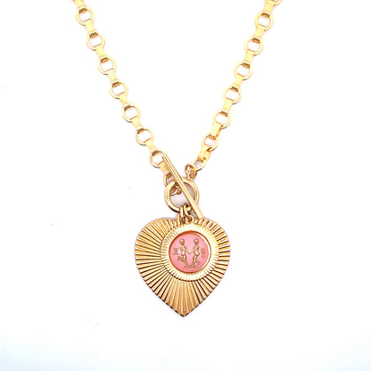 A gold ribbed heart-shaped pendant necklace with a Gemini zodiac twins design in pink enamel center with toggle clasp closure on a gold round link chain against a white background