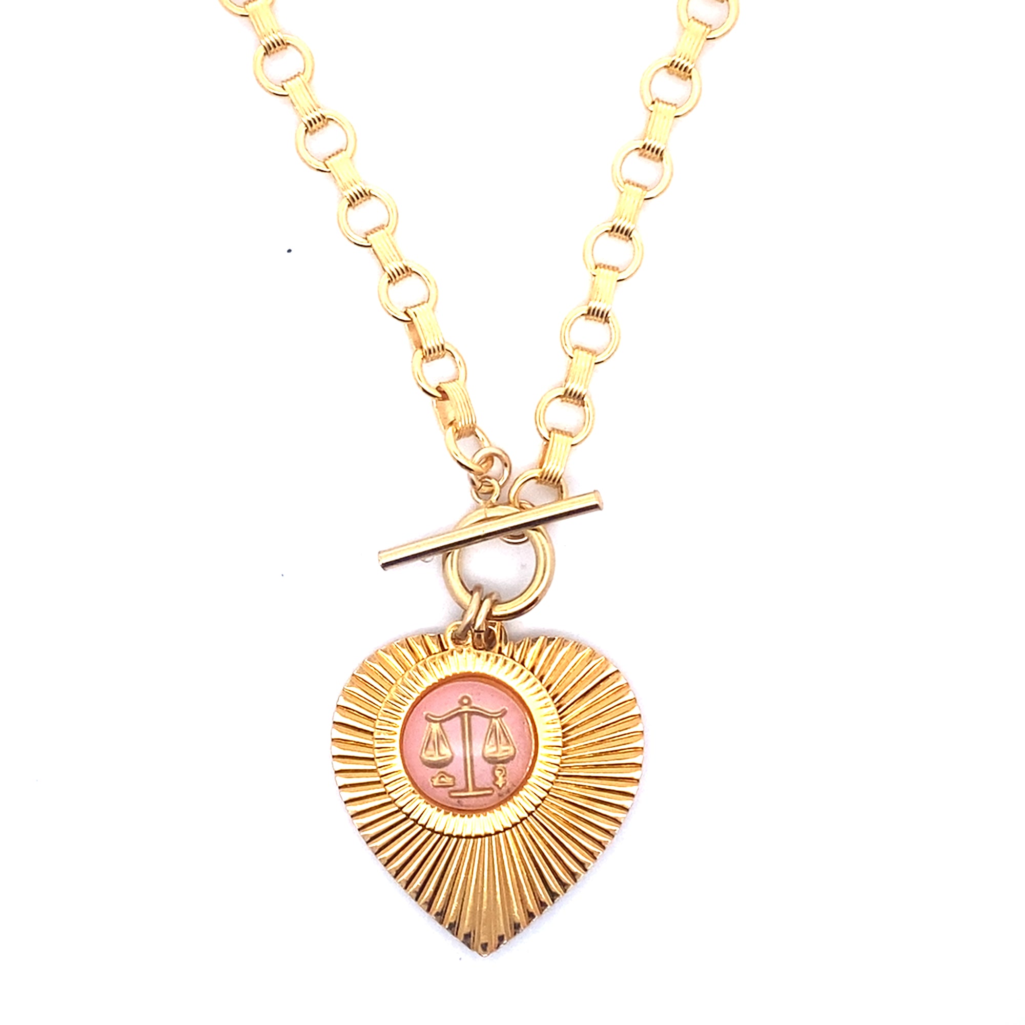A gold ribbed heart-shaped pendant necklace with a Libra zodiac scales design in pink enamel center with toggle clasp closure on a gold round link chain against a white background