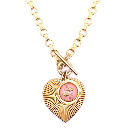 A gold ribbed heart-shaped pendant necklace with a Pisces zodiac fish design in pink enamel center with toggle clasp closure on a gold round link chain against a white background