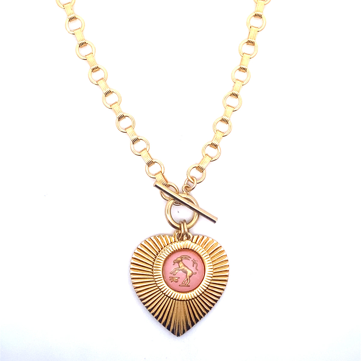 A gold ribbed heart-shaped pendant necklace with a Capricorn zodiac goat design in pink enamel center with toggle clasp closure on a gold round link chain against a white background
