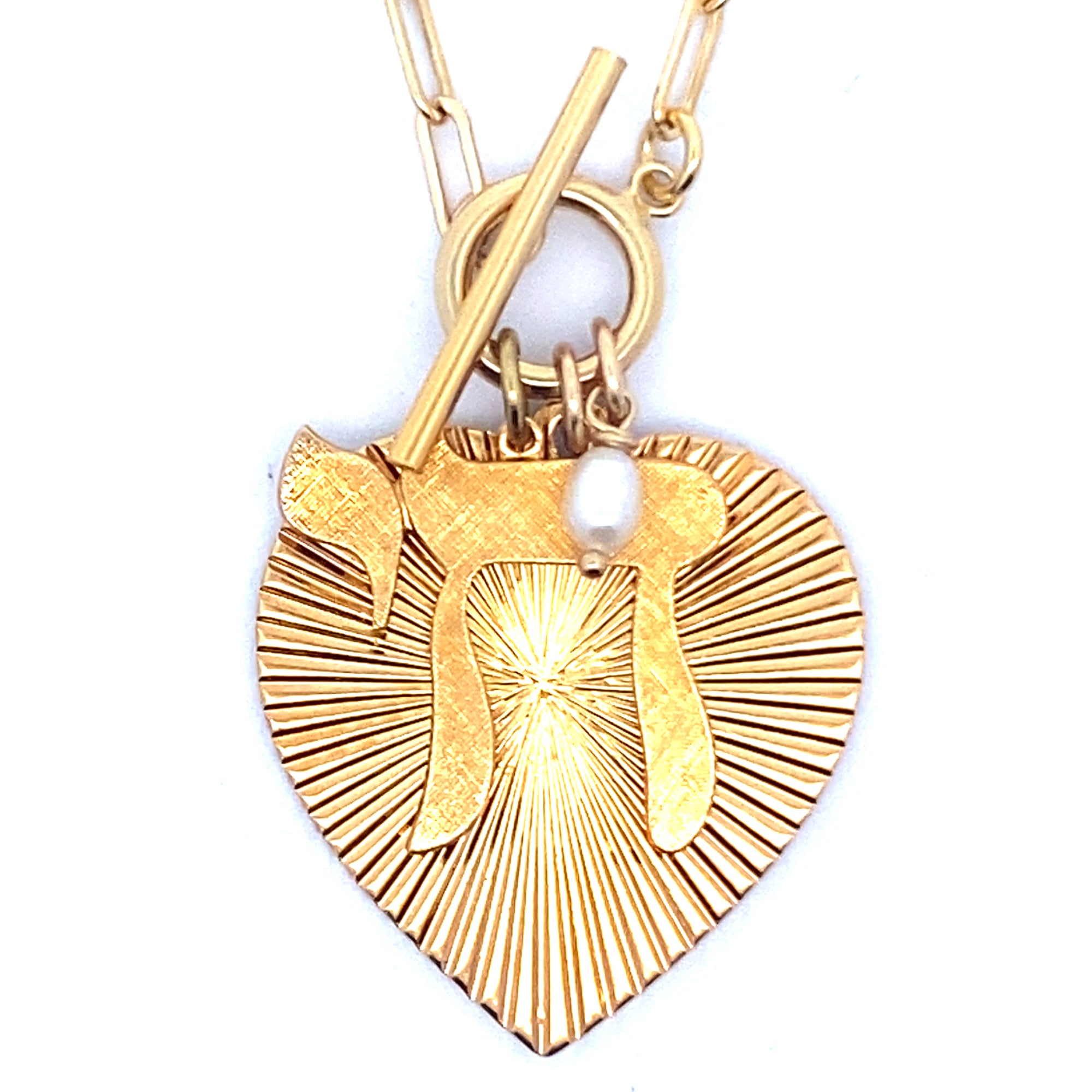 Close-up of the back of a gold ribbed heart-shaped pendant with starburst pattern and small pearl charm on a gold paperclip chain with toggle clasp against a white background