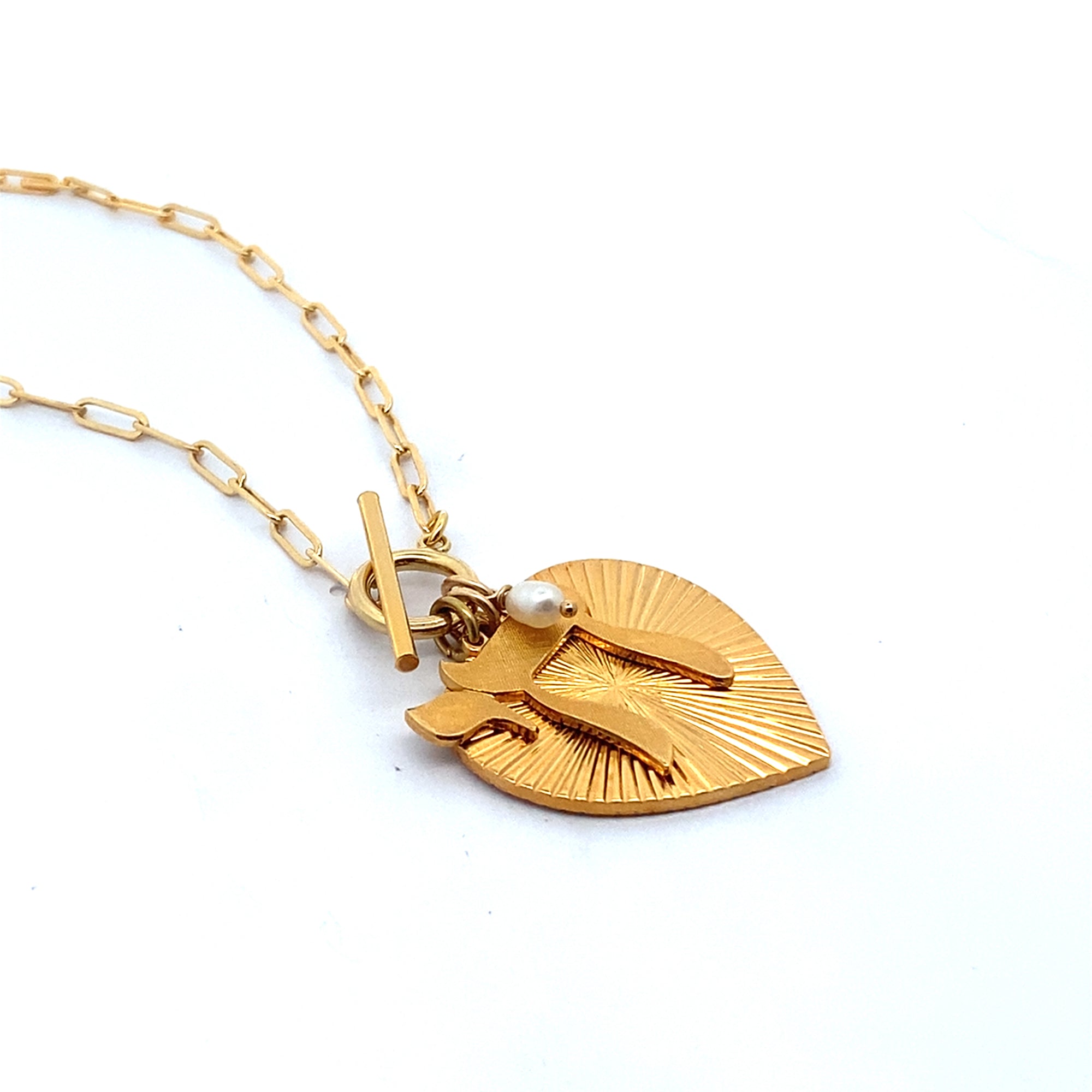 Side angle view of the back of a gold ribbed heart-shaped pendant with starburst pattern and small pearl charm on a gold paperclip chain with toggle clasp against a white background