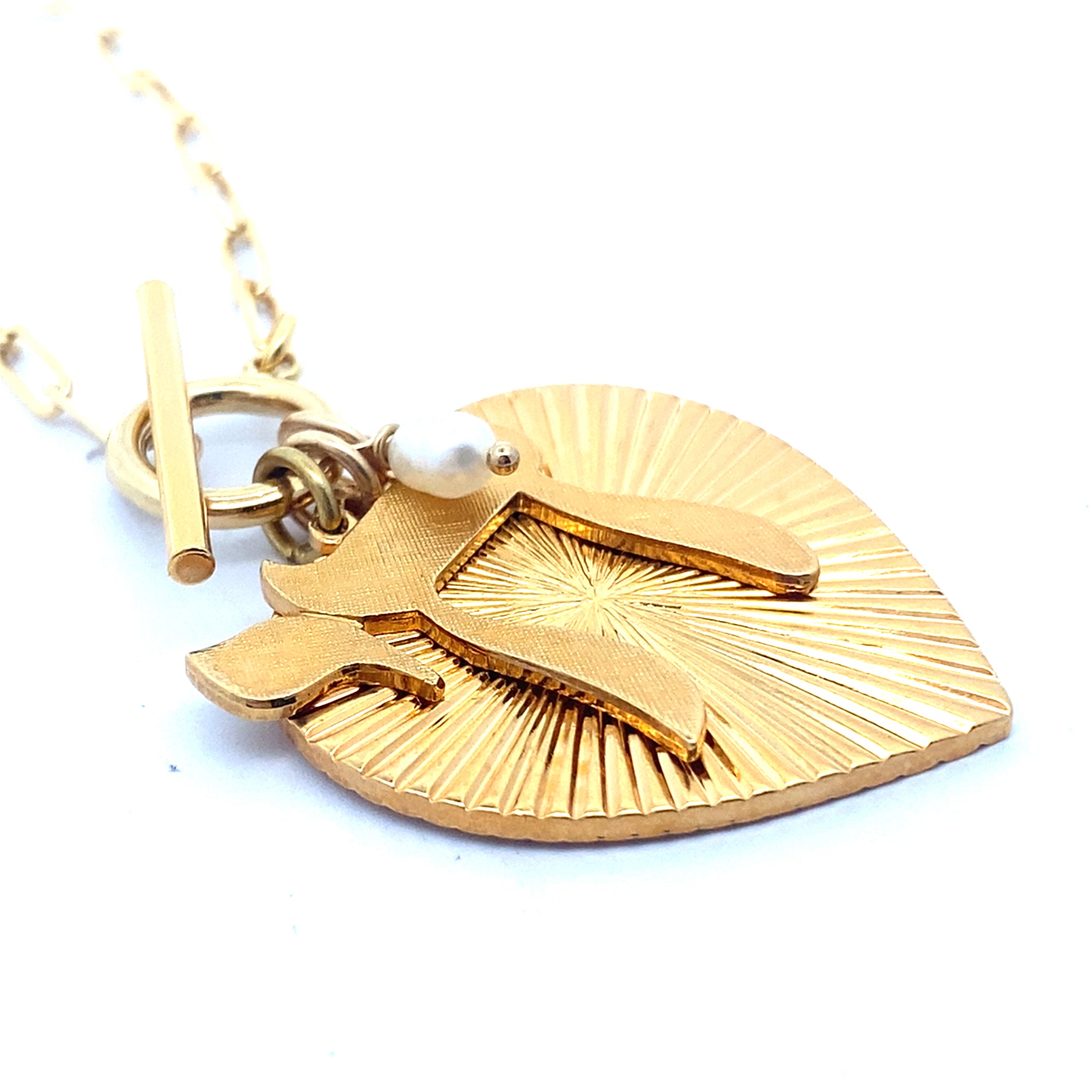 Close-up angled view of the back of a gold ribbed heart-shaped pendant with starburst pattern and small pearl charm on a gold paperclip chain with toggle clasp against a white background
