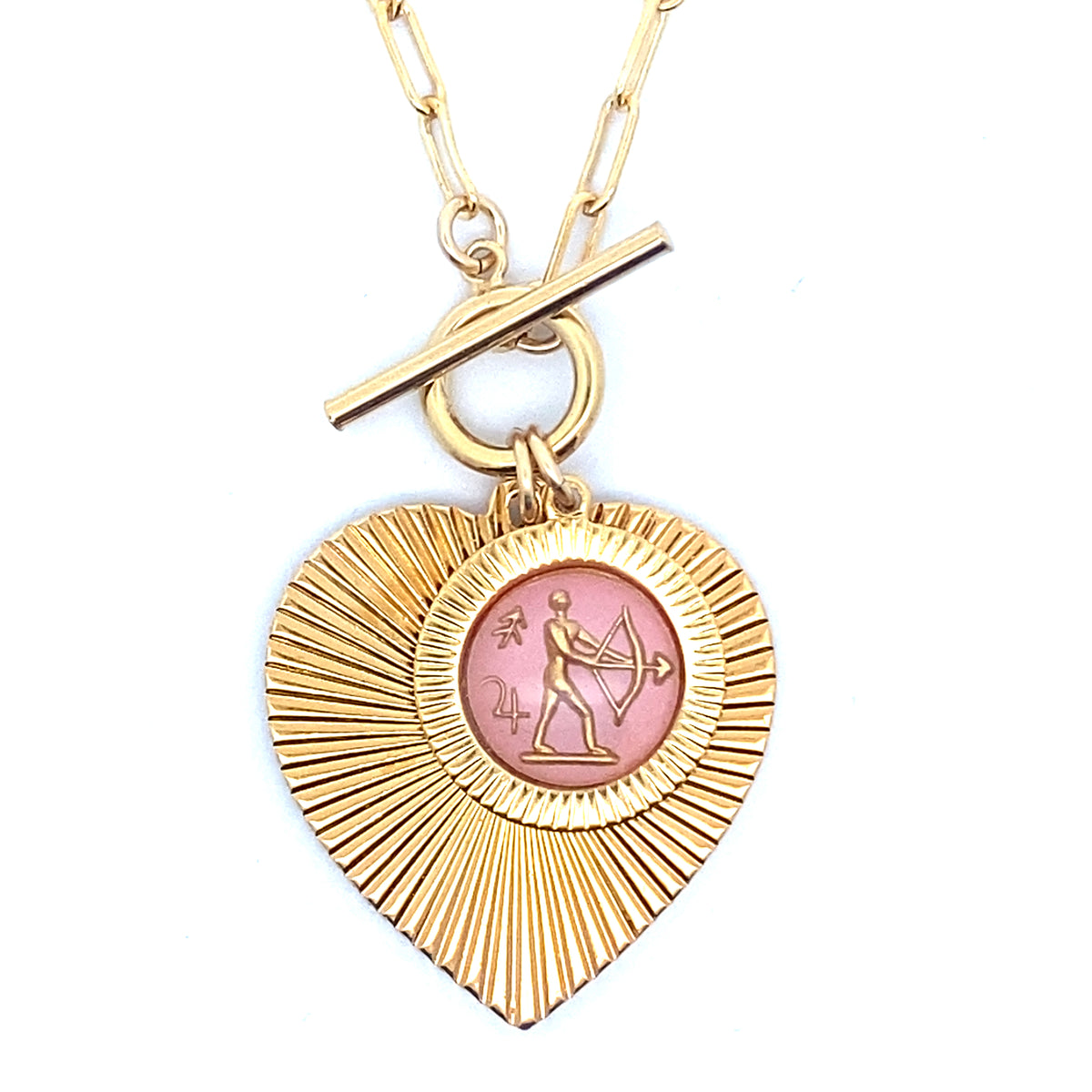 Close-up of a gold ribbed heart-shaped pendant necklace with a Sagittarius zodiac archer design in pink enamel center with toggle clasp closure on a gold paperclip chain worn on a person in a white top
