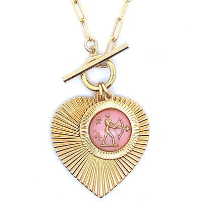 Close-up of a gold ribbed heart-shaped pendant necklace with a Sagittarius zodiac archer design in pink enamel center with toggle clasp closure on a gold paperclip chain worn on a person in a white top