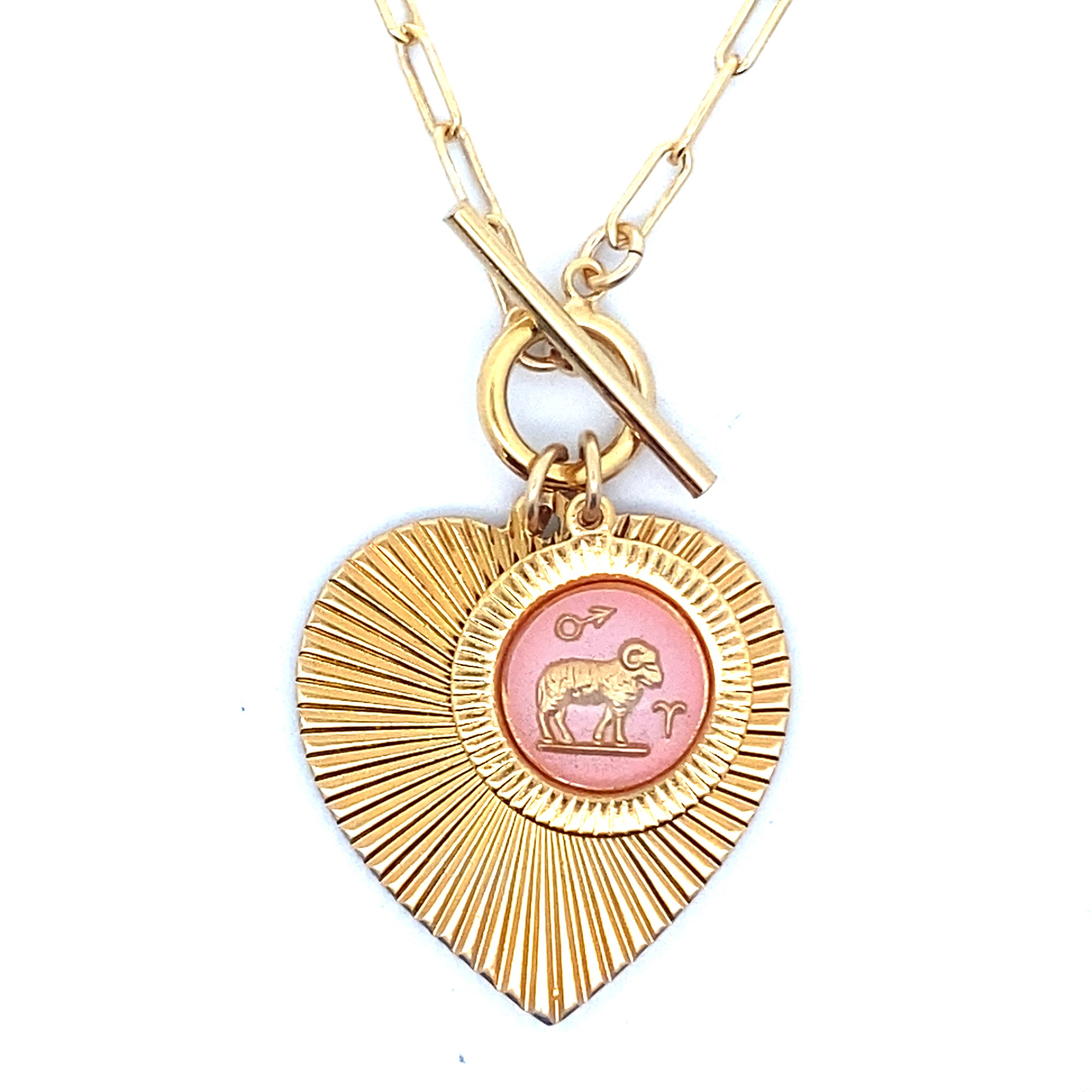 Close-up of a gold ribbed heart-shaped pendant necklace with an Aries zodiac ram design in pink enamel center with toggle clasp closure on a gold paperclip chain worn on a person in a white top