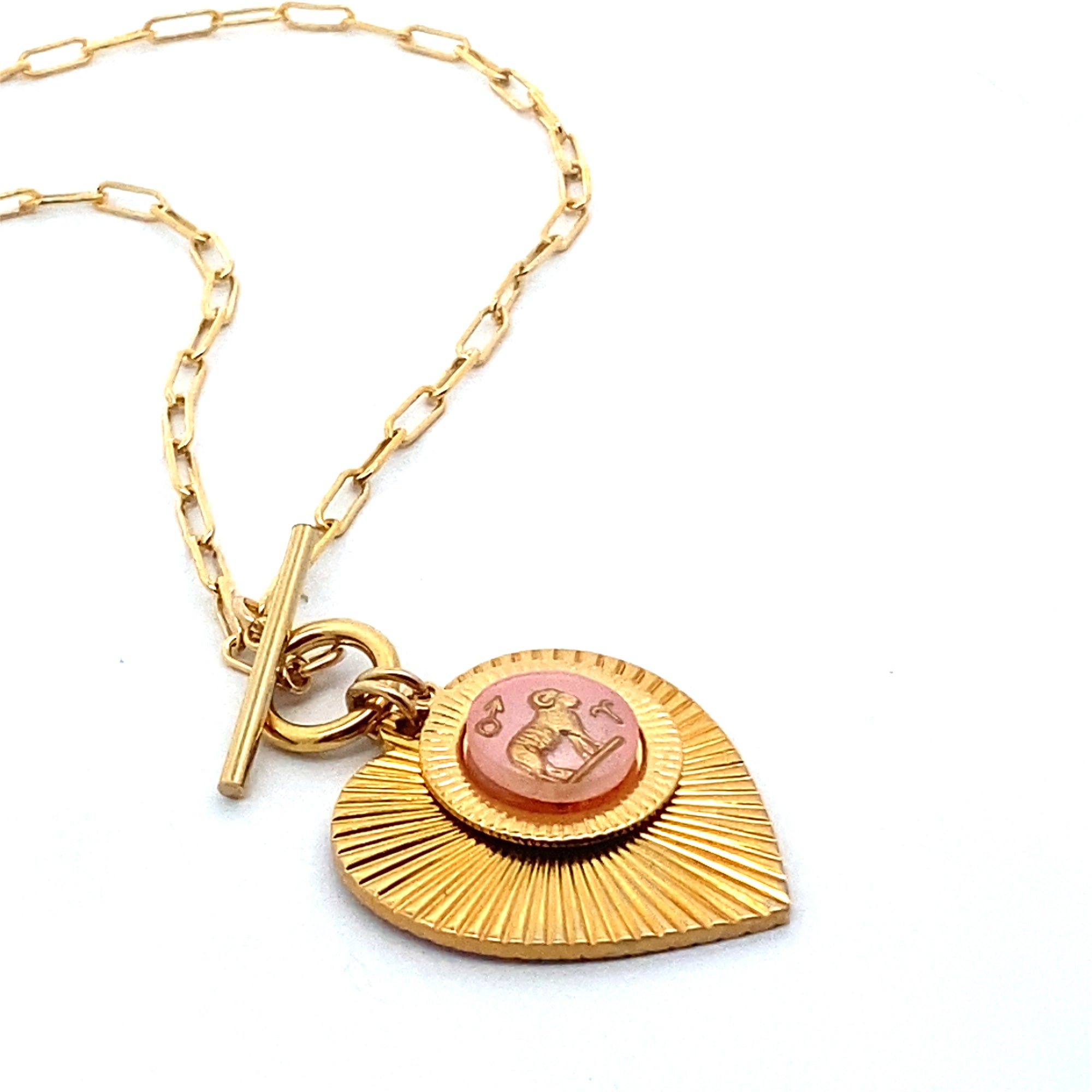 A gold ribbed heart-shaped pendant necklace with an Aquarius zodiac water bearer design in pink enamel center with toggle clasp closure on a gold paperclip chain against a white background