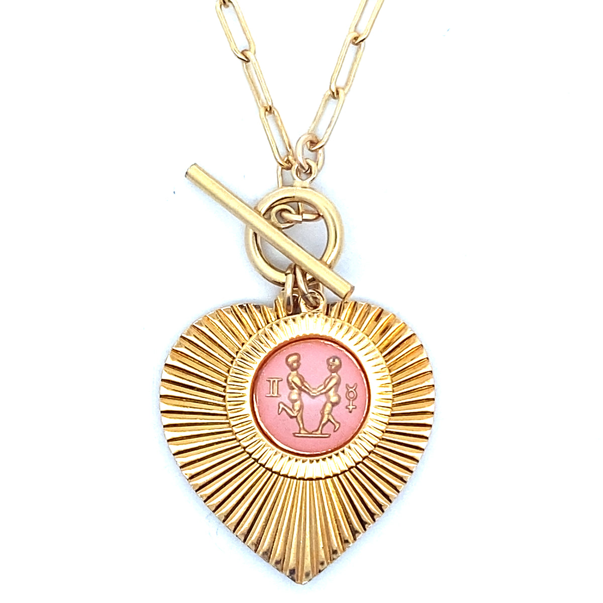 Close-up of a gold ribbed heart-shaped pendant necklace with a Gemini zodiac twins design in pink enamel center with toggle clasp closure on a gold paperclip chain worn on a person in a white top