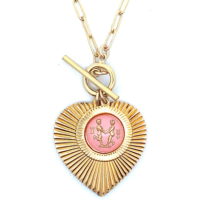 Close-up of a gold ribbed heart-shaped pendant necklace with a Gemini zodiac twins design in pink enamel center with toggle clasp closure on a gold paperclip chain worn on a person in a white top