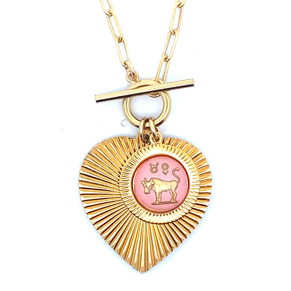 Close-up of a gold ribbed heart-shaped pendant necklace with a Taurus zodiac bull design in pink enamel center with toggle clasp closure on a gold paperclip chain worn on a person in a white top