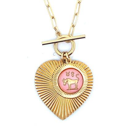Close-up of a gold ribbed heart-shaped pendant necklace with a Taurus zodiac bull design in pink enamel center with toggle clasp closure on a gold paperclip chain worn on a person in a white top