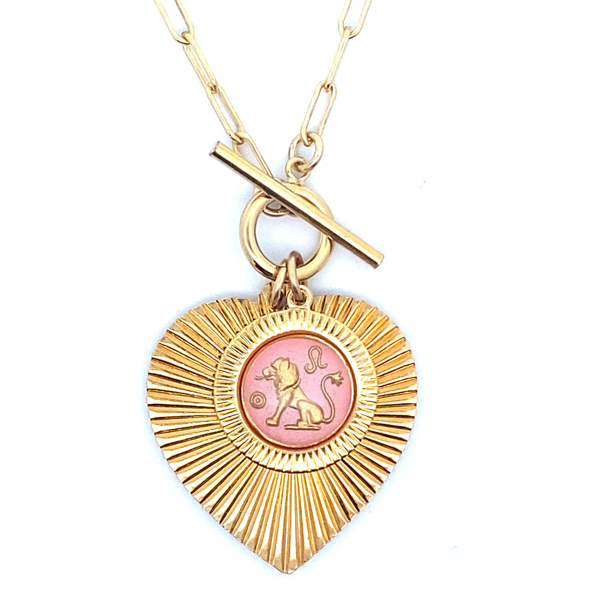 A gold ribbed heart-shaped pendant necklace with a Leo zodiac lion design in pink enamel center with toggle clasp closure on a gold paperclip chain against a white background