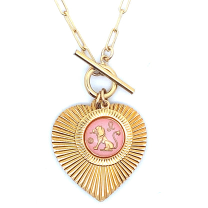 A gold ribbed heart-shaped pendant necklace with a Leo zodiac lion design in pink enamel center with toggle clasp closure on a gold paperclip chain against a white background